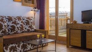 A cozy room with a cow print sofa, a small table with two glasses of orange juice, a TV on a wooden cabinet, and a sliding glass door leading to a balcony with a wooden railing and a view of snow-capped mountains. - 150m from ski slopes, swimming pool, balcony, tv, ski locker, 50-55m², Montchavin La Plagne (Bellentre)