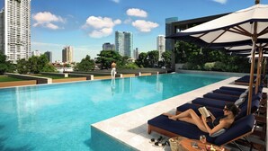 Zona de piscina con sillones de descanso y sombrillas, una piscina azul cristalina y un horizonte urbano al fondo.