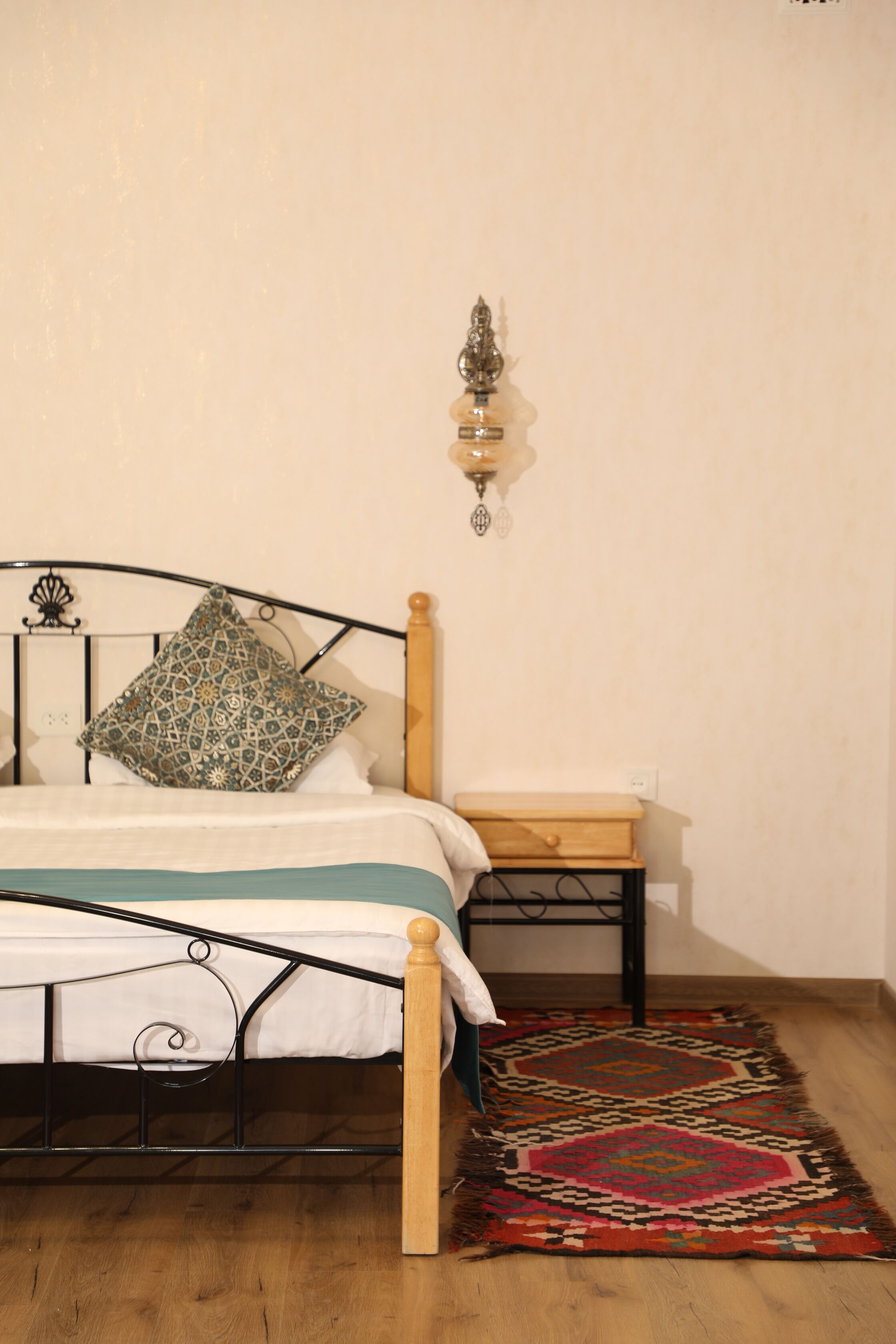 A bed with a patterned pillow, a bedside table, a wall-mounted light fixture, and a patterned rug on a wooden floor.