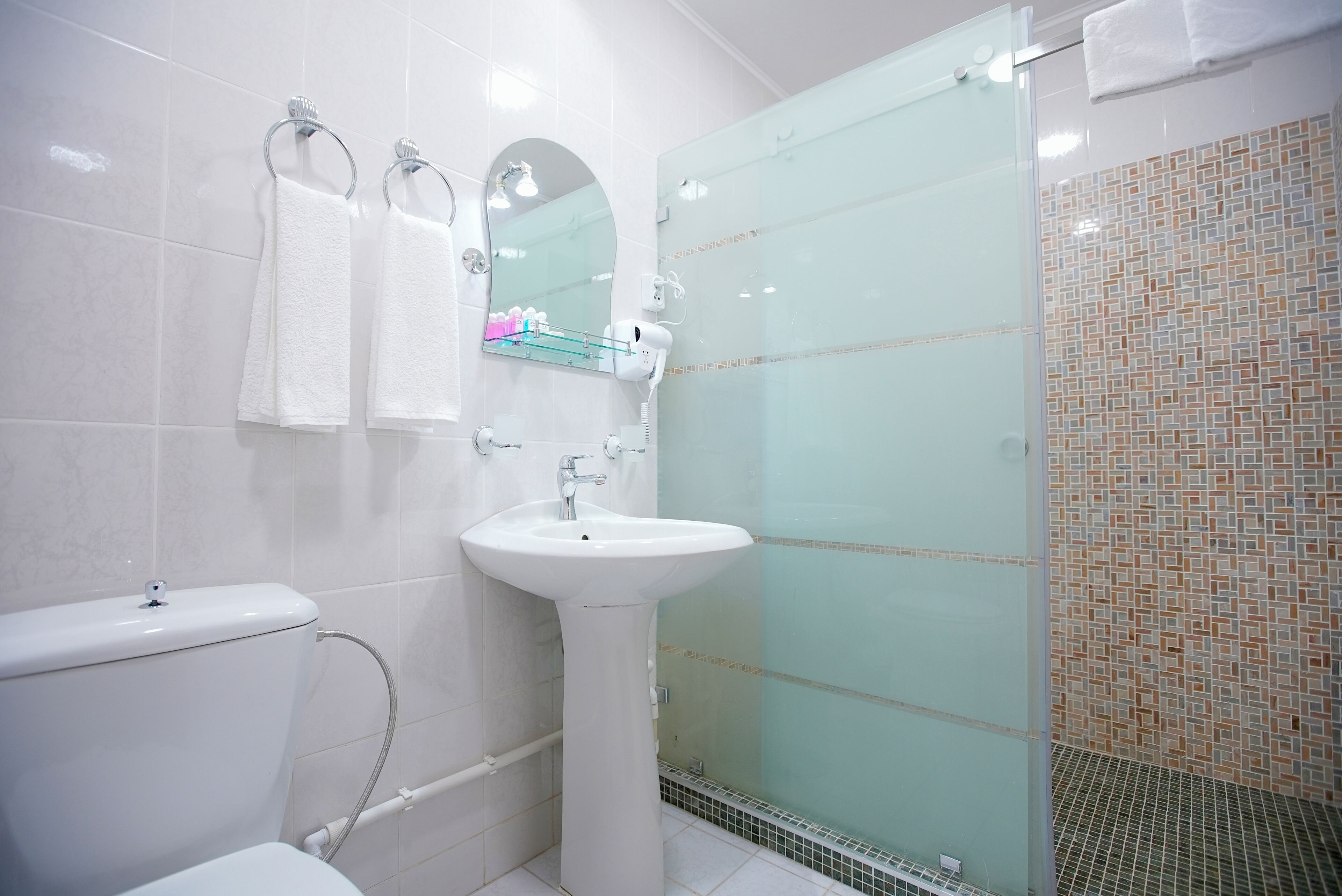 Une salle de bain moderne avec des toilettes blanches, un lavabo sur colonne, une cabine de douche en verre et un mur de douche recouvert de carreaux.