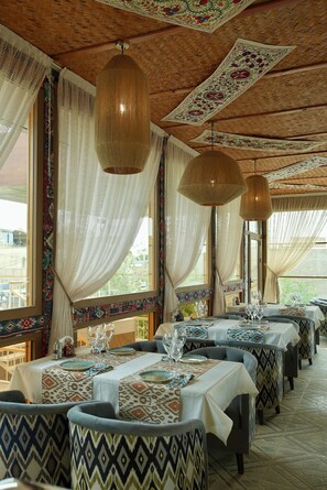 Un restaurante con mesas cubiertas por manteles estampados, lámparas tejidas colgantes y cortinas drapeadas.