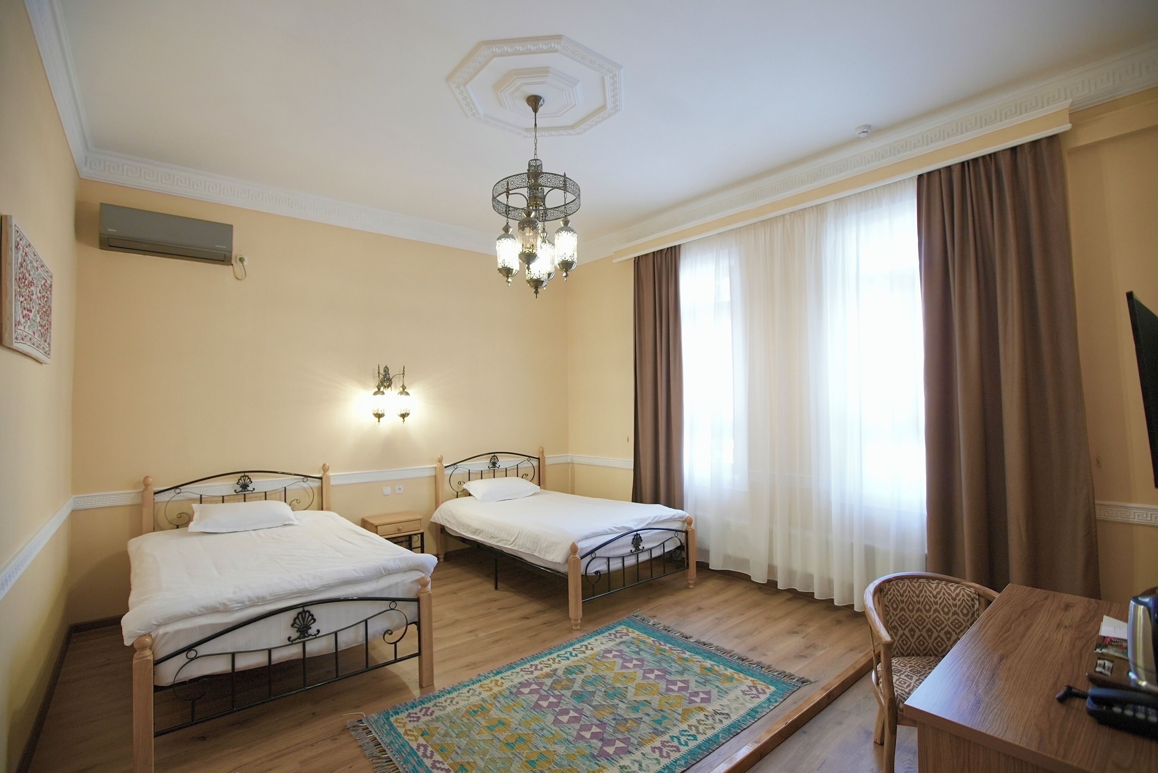 Habitación de hotel con dos camas, piso de madera, alfombra estampada, lámpara de araña y escritorio con una silla.