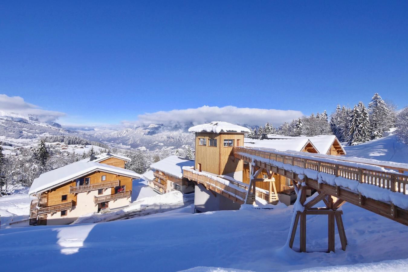 A wooden chalet with a balcony, a wooden bridge, and a snow-covered mountain backdrop.