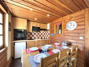 A compact kitchen with wooden cabinets, a microwave, and a small refrigerator. A dining table with chairs is set for two, and a wall clock is visible.