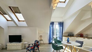 A spacious living area with a flat-screen TV, a floral chair, and a dining table with chairs. There are skylights and a kitchen area visible. - Apartament Mundo Kołobrzeg - 365PAM (Kolobrzeg)
