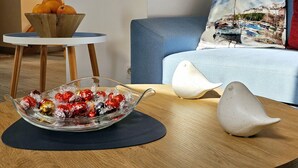 A living room with a round wooden coffee table, a glass bowl with wrapped chocolates, a white decorative bird, a blue sofa, and a small white side table with a bowl of oranges. - Apartament Mundo Kołobrzeg - 365PAM (Kolobrzeg)