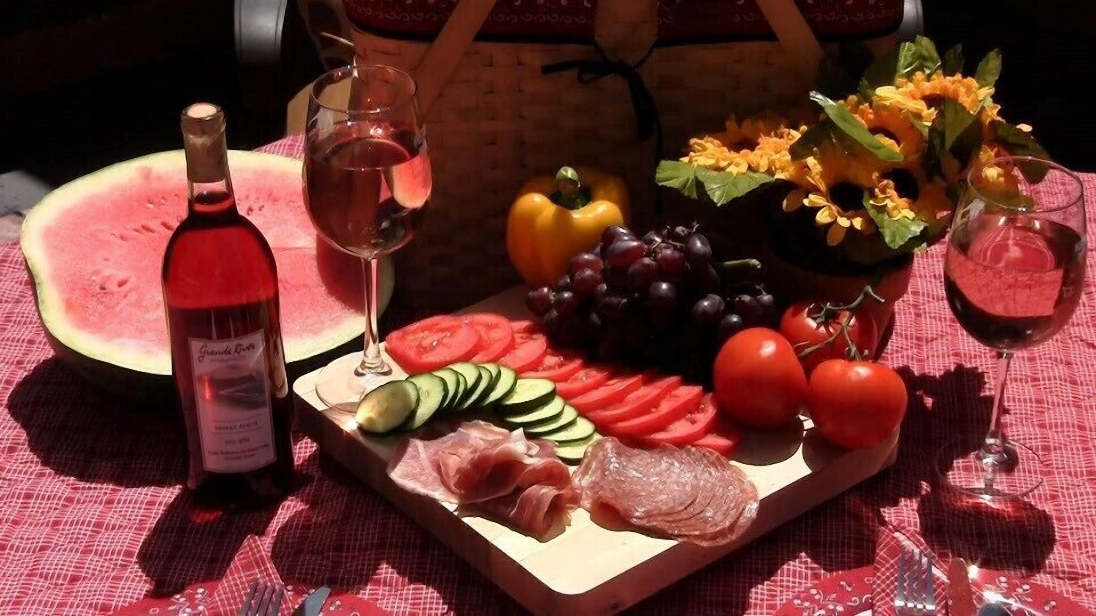 Un montaje para picnic con sandía, uvas, tomates, pepinos, jamón serrano y una botella de vino sobre un mantel a cuadros.