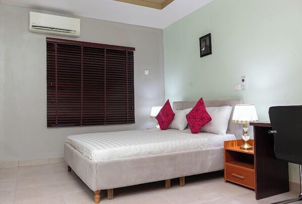 A bedroom with a bed, a nightstand, a lamp, a chair, a window with blinds, and an air conditioning unit.