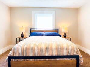 Un lit bien fait, recouvert d’une couvre-lit à rayures, avec deux lampes de chevet et une fenêtre équipée de stores.