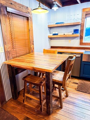 Une table à manger en bois avec des chaises, un luminaire suspendu et un coin cuisine avec une étagère et un tableau encadré.