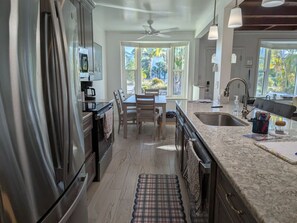 A modern kitchen with a stainless steel refrigerator, a dining area with a table and chairs, a large window with a view of greenery, and a ceiling fan.