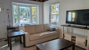 Ein Wohnzimmer mit einem beigefarbenen Sofa, einem Holztisch, einem Fernseher auf einem Standfuß und einem Fenster mit Blick auf Palmen.