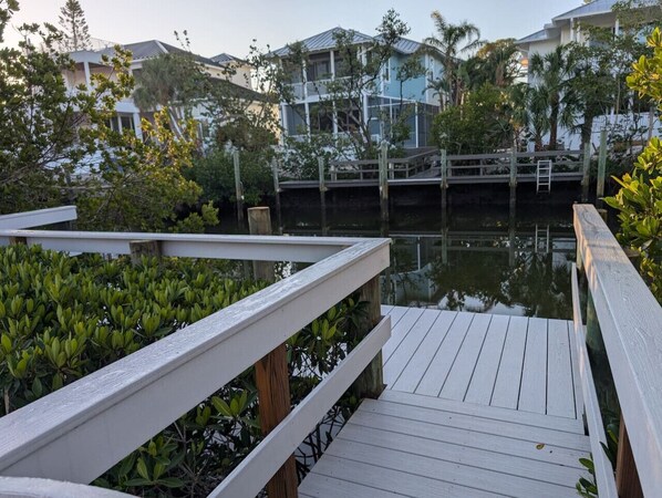 A wooden dock leading to a calm water body, surrounded by lush greenery and residential buildings in the background.