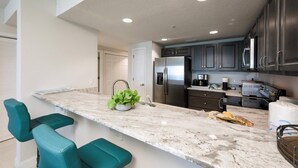 A modern kitchen with dark cabinets, a marble countertop, and teal bar stools. - Unit 203 Aruba - 3 Bedroom Ocean Front (Daytona Beach Shores)