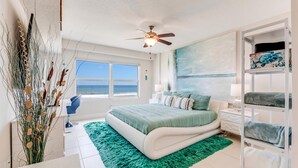 A bedroom with a bed, a green rug, a ceiling fan, a window with a view of the ocean, and a shelf with towels. - Unit 203 Aruba - 3 Bedroom Ocean Front (Daytona Beach Shores)