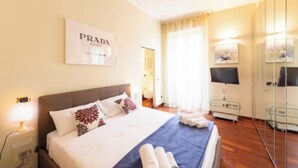 Quarto de hotel com uma cama, uma TV, um espelho e um letreiro Prada na parede.