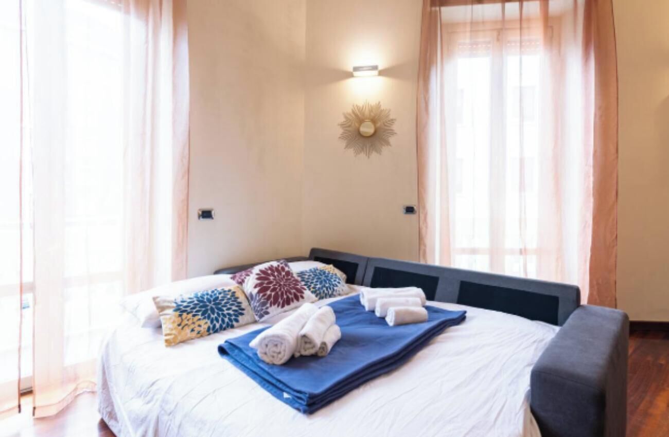 Een net opgemaakt bed met kussens en handdoeken, een wandlamp en een raam met dunne gordijnen.