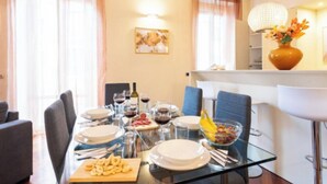 Uma área de refeições com mesa de vidro posta para uma refeição, incluindo pratos, talheres e uma tigela de frutas.