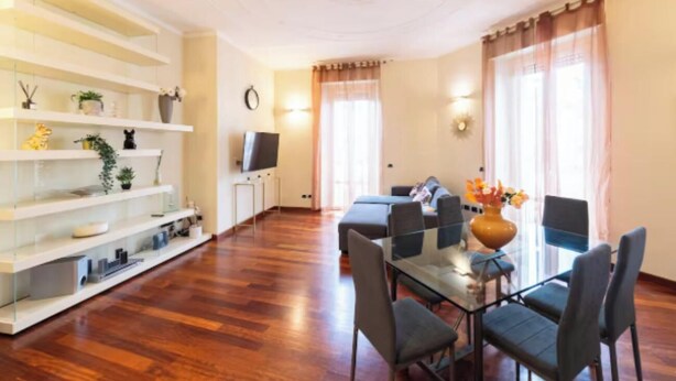 Sala de estar moderna com área de jantar, piso de madeira, estantes embutidas, televisão de tela plana e uma janela grande com cortinas.