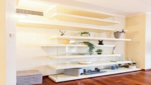 Um interior moderno com prateleiras brancas suspensas, um banco cinzento e piso de madeira.