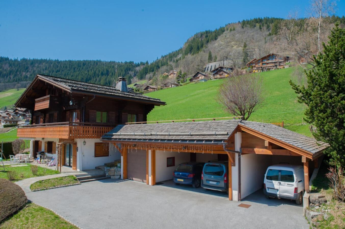 A wooden chalet with a balcony, a covered garage, and a driveway with two cars parked.