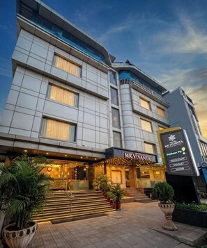 Un hôtel à plusieurs étages doté d’une façade moderne, d’une entrée imposante avec des marches et d’une signalétique indiquant les différents services proposés, tels qu’un restaurant, un bar et des chambres.