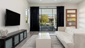 A modern living room with a flat-screen TV, a glass-top coffee table, a white sofa, a dark wooden console, and a sliding glass door leading to an outdoor area.