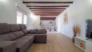 Sala de estar com sofá seccional, televisão em um móvel, piso de madeira e cozinha ao fundo.