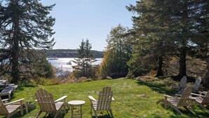 A view of a lakeside deck with Adirondack chairs and a small table, surrounded by trees and overlooking a body of water.
