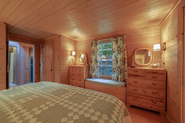 A wooden cabin bedroom with a bed, dresser, and window with curtains.