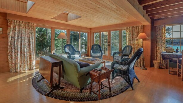 A cozy cabin interior with a wooden ceiling, large windows, and a round table with chairs.