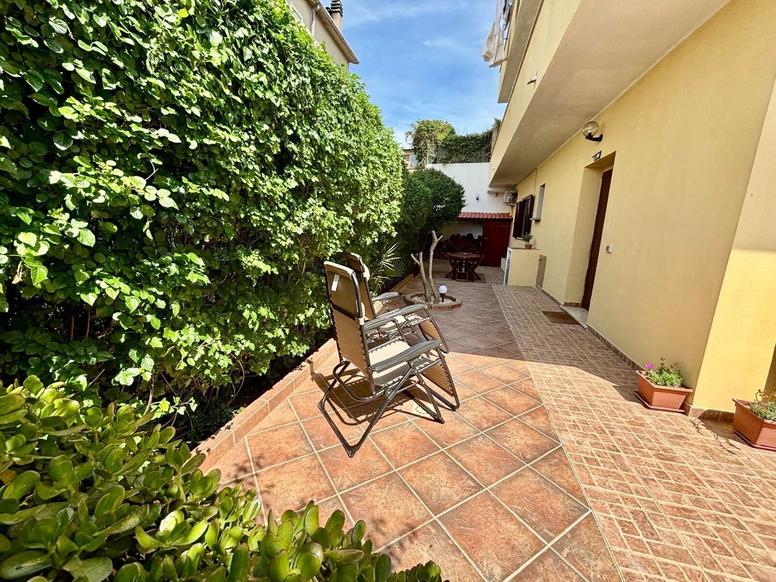 Une terrasse avec une table et des chaises, entourée d’une haie et d’un bâtiment.