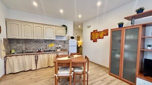 A compact kitchen with wooden cabinets, a dining table with chairs, and a cabinet with glass doors. - Cozy apartment, 10 min. from Alghero, ideal for families (Olmedo)