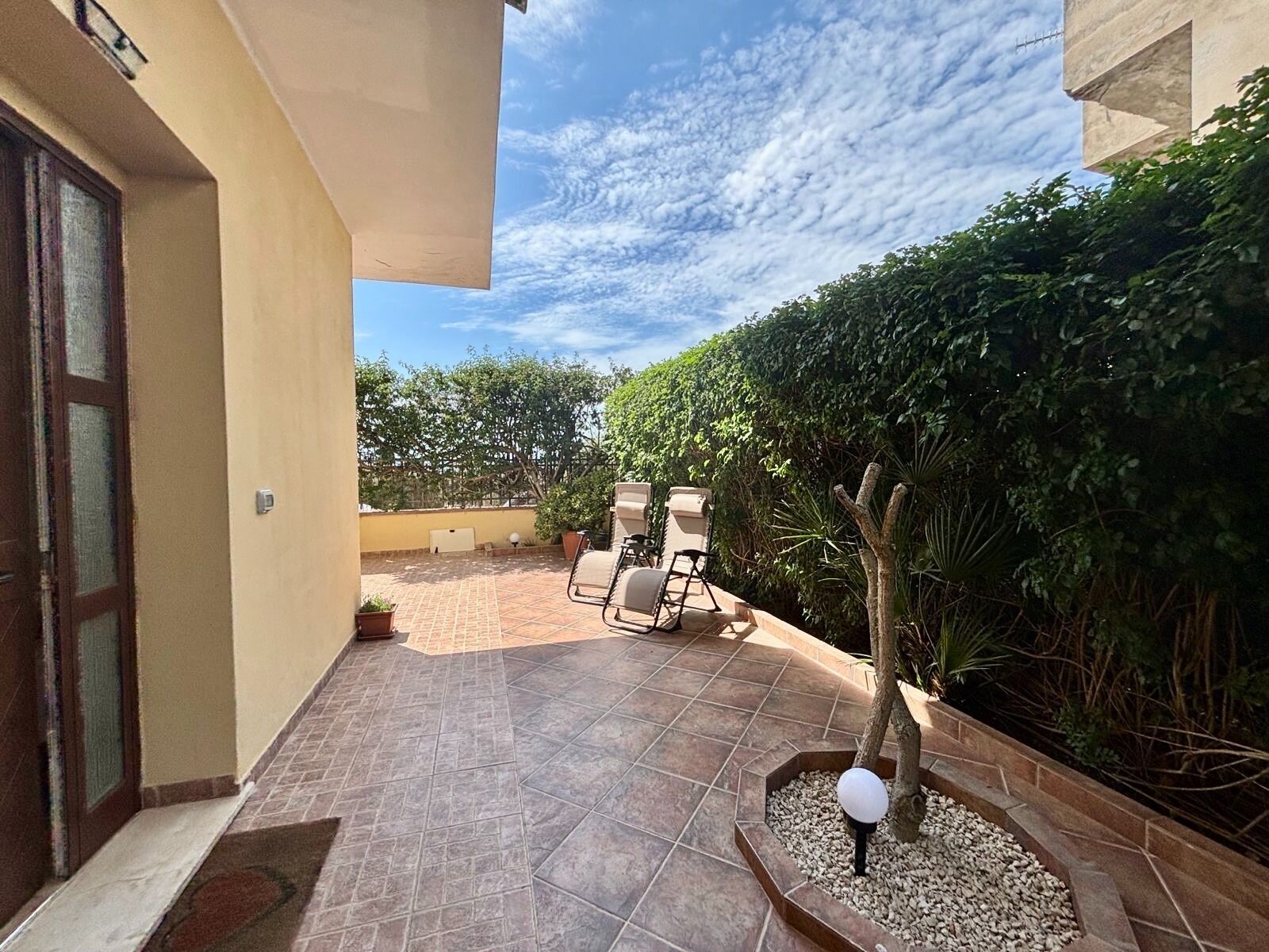 Un'area patio con pavimento piastrellato, mobili da esterno e un piccolo giardino con un lampione.