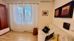 A room with a wooden floor, a white sofa with decorative pillows, a wooden cabinet, a window with patterned curtains, and a wall-mounted air conditioner. - Cozy apartment, 10 min. from Alghero, ideal for families (Olmedo)