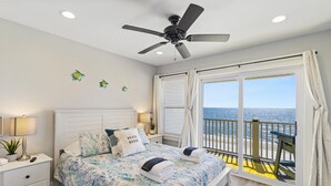 A bedroom with a bed, a ceiling fan, a nightstand, a lamp, a window with curtains, and a view of the ocean. - Gulf-front! Beachfront! Directly on the Sand! New Boardwalk! Amazing Ocean View! (Mexico Beach)