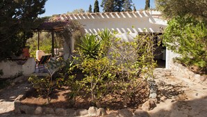 Un edificio rústico con fachada de piedra, un área de estar al aire libre cubierta con sillas y un jardín con diversas plantas.