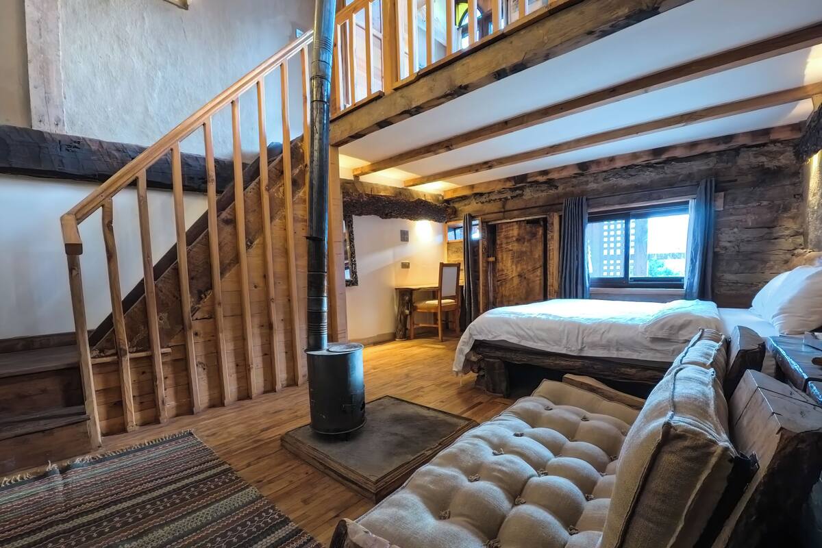 A rustic room with a wooden staircase, a bed with a white duvet, a sofa, and a large window.