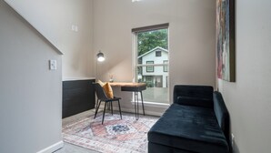 Una habitación pequeña y moderna con un sofá negro, un escritorio con una silla, un ventanal grande y una alfombra estampada.