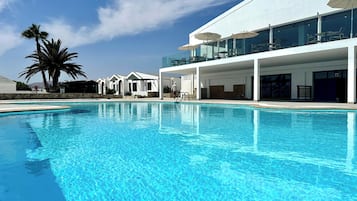 A clear blue pool with a modern building in the background, featuring outdoor seating and a covered area.