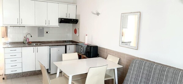 A compact kitchen with white cabinets, a sink, a small refrigerator, and a dining area with a table and chairs. - Caleta de Fuste Apartment- Sleeps4- Pool- Garden (Antigua)