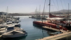 Um porto com vários barcos atracados, céu claro e paisagem montanhosa ao fundo.