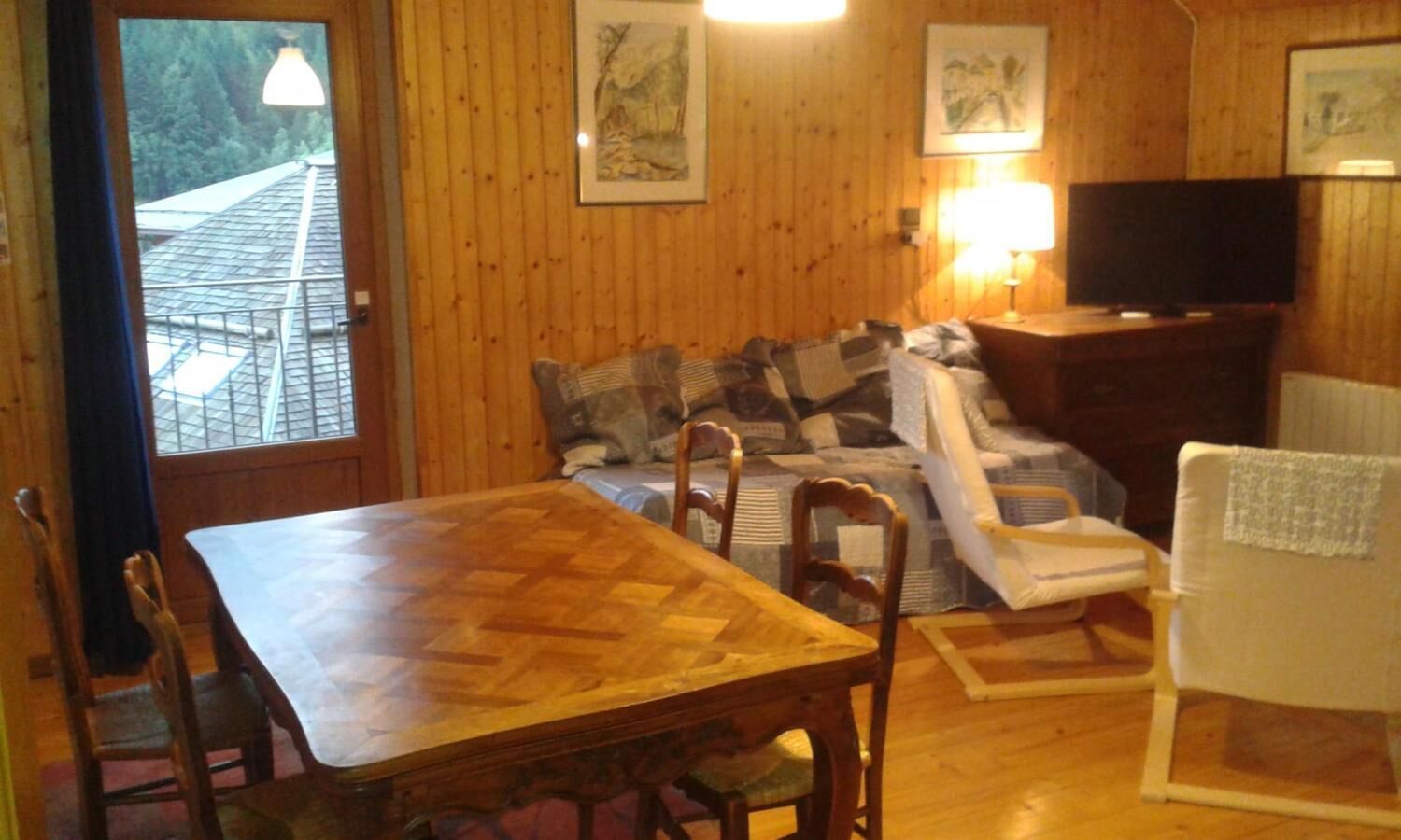 Et hyggeligt værelse med et træspisebord, en sofa med mønstrede puder, et fjernsyn og et vindue med udsigt over et tag.