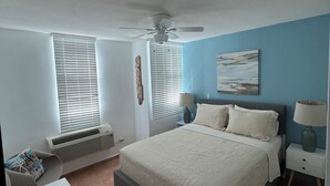 A bedroom with a bed, a ceiling fan, two bedside tables with lamps, a wall-mounted artwork, and a window with blinds.