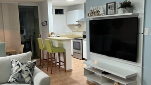 A modern living room with a grey sofa, a TV on a white stand, and a kitchen in the background.