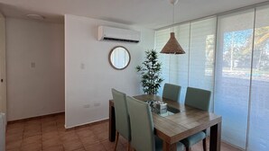 A dining area with a wooden table, four chairs, a hanging light fixture, a potted plant, and a large window with a view.