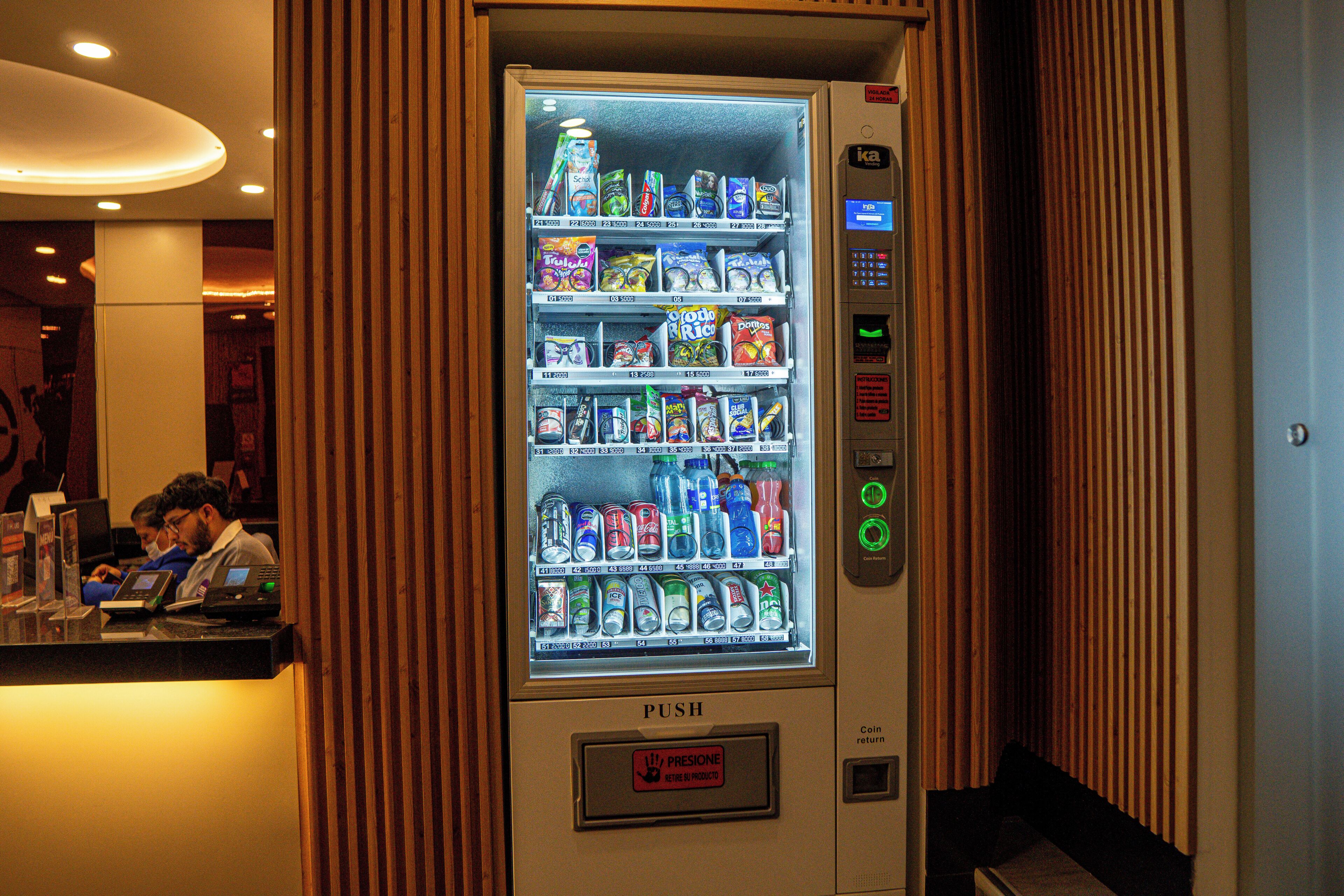 Máquina automática com variedade de lanches e bebidas, interface de pagamento e um botão.