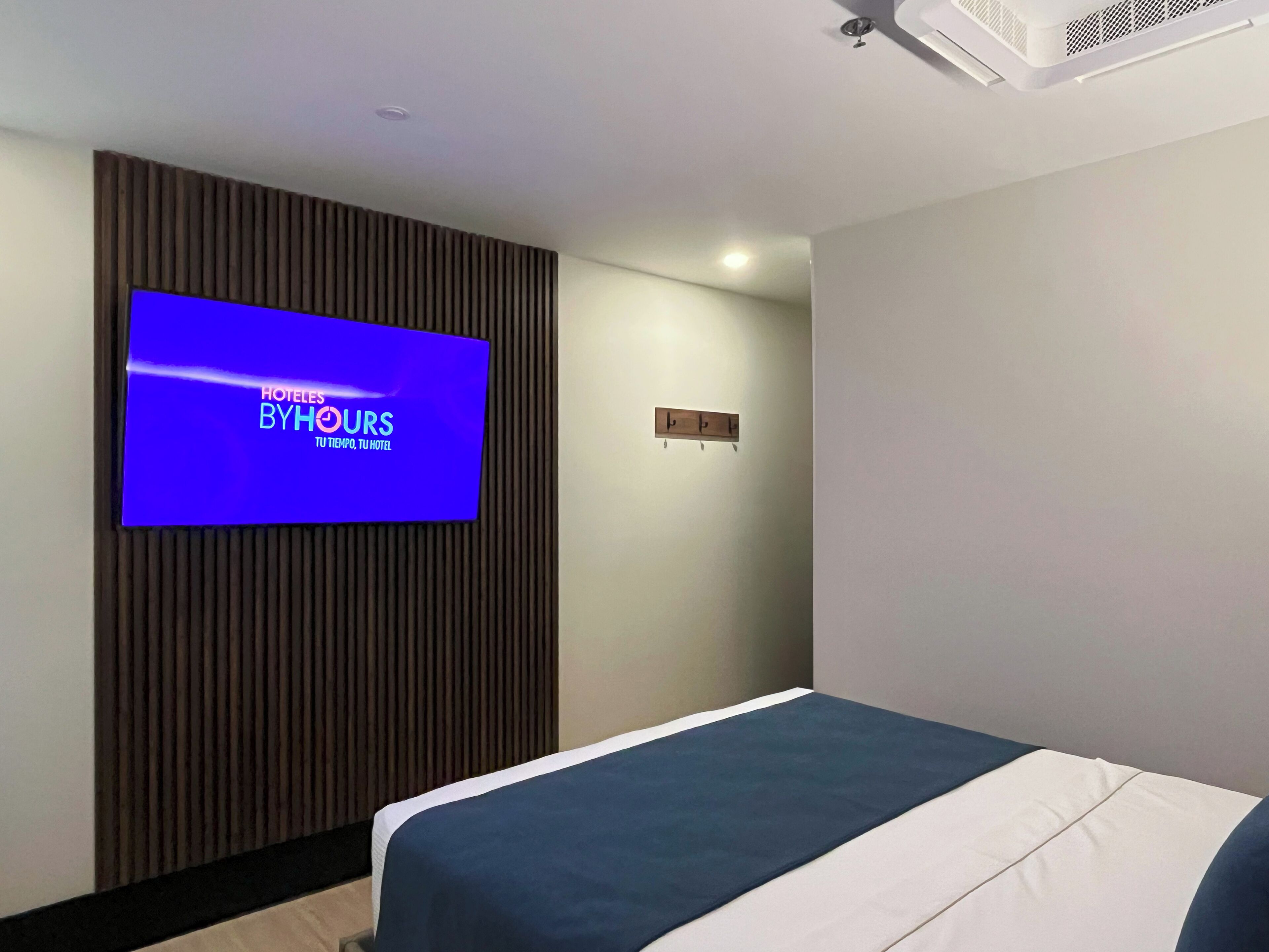 A hotel room with a bed, a flat-screen TV mounted on a wooden panel, and a small wooden shelf with a clock.