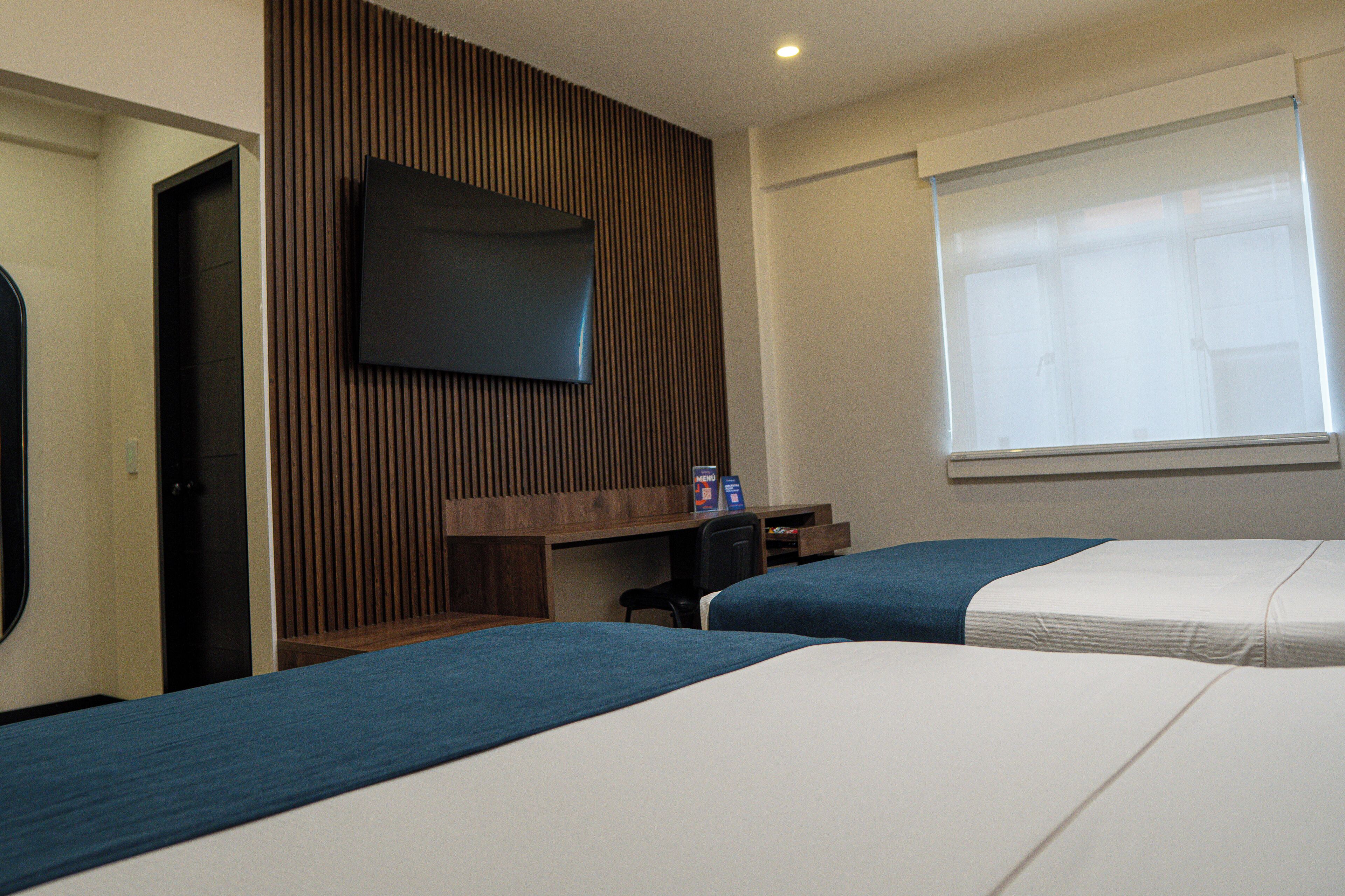 Une chambre d’hôtel avec deux lits, un téléviseur fixé au mur, un bureau et une fenêtre avec des stores.