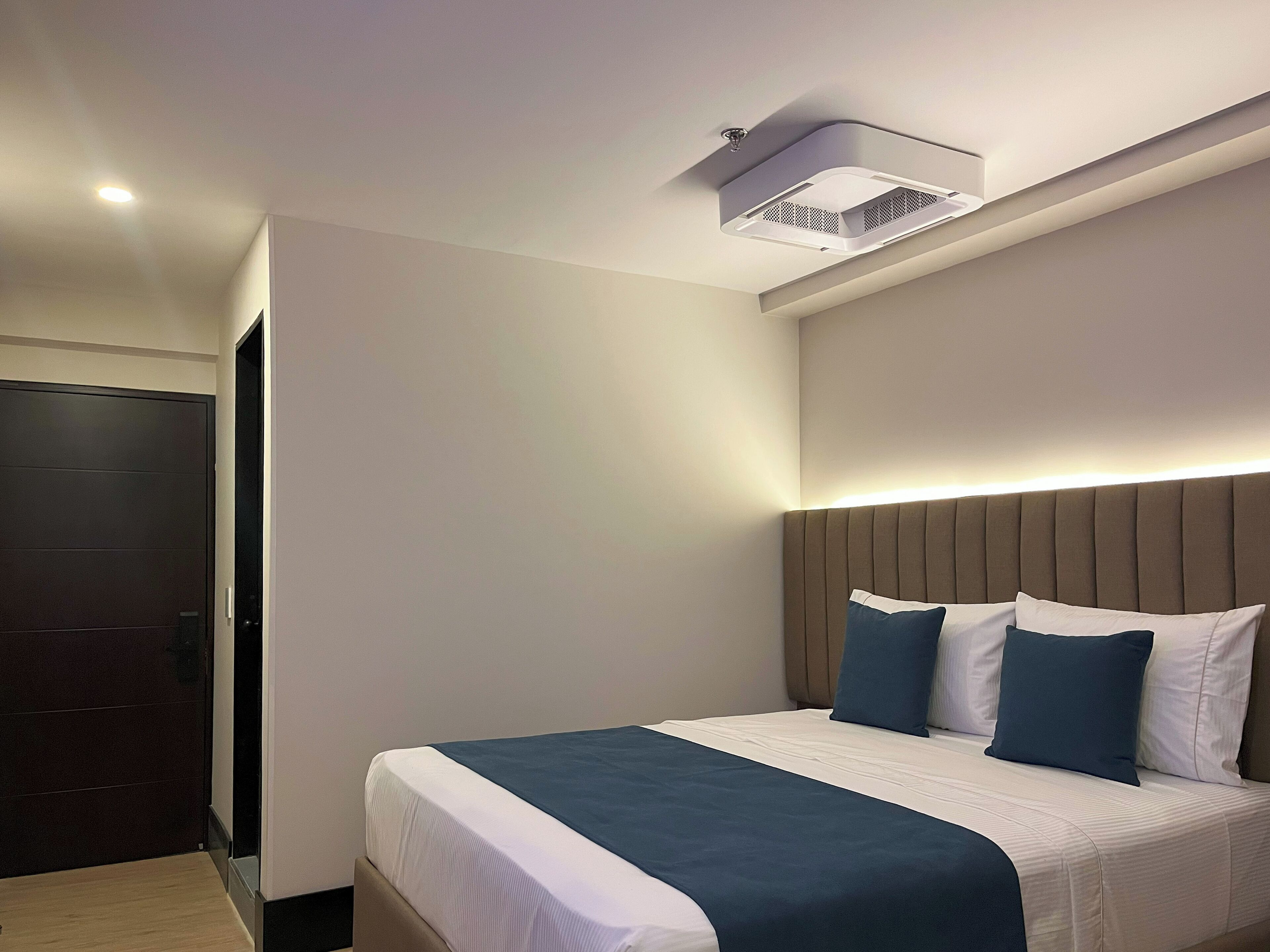 A modern hotel room with a large bed, a headboard with vertical lines, and a ceiling-mounted air conditioning unit.